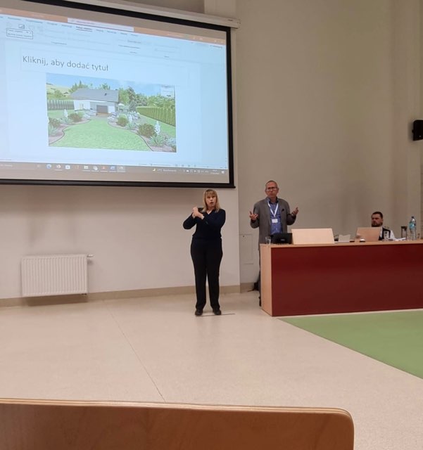 Na zdjęciu prowadzący konferencje oraz tłumacz stojący przy wyświetlanej prezentacji na ścianie 